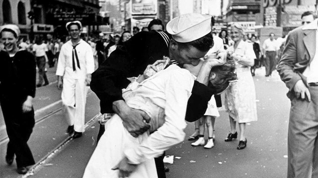 A historic black and white photo depicting a sailor passionately kissing a woman in a white dress on a busy street, surrounded by crowds celebrating, capturing a moment of love and joy.