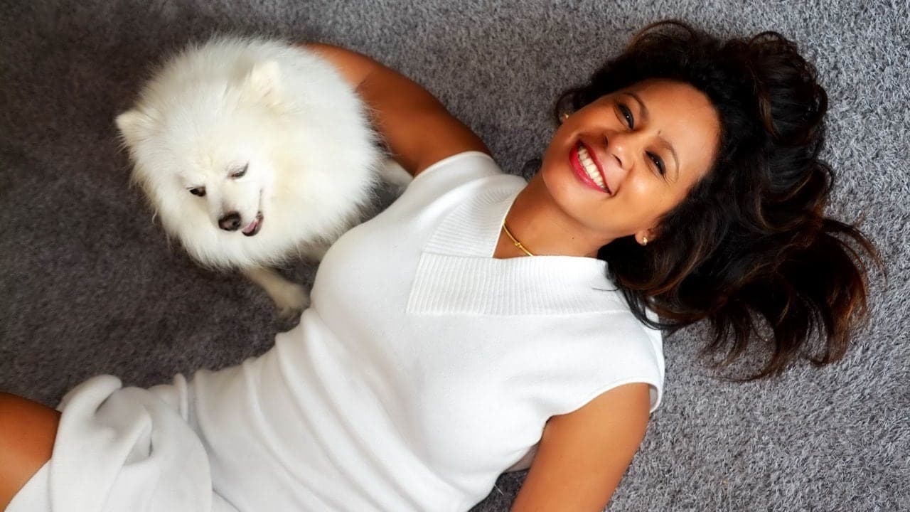 A woman lying on a gray carpet, smiling while interacting with a fluffy white dog beside her.