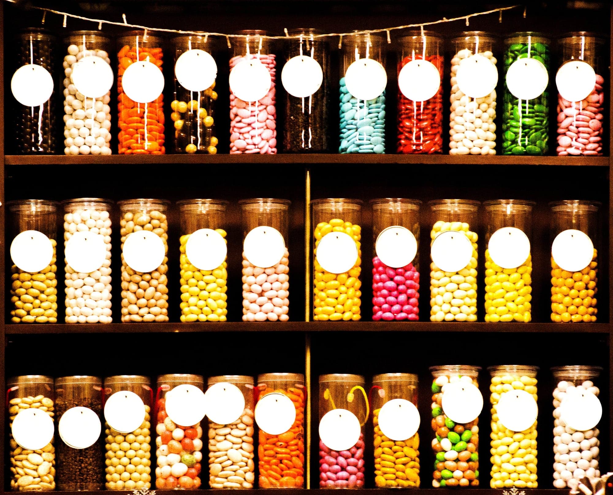 A colorful display of assorted candy jars on a shelf, each filled with different types and colors of candies, labeled with tags.