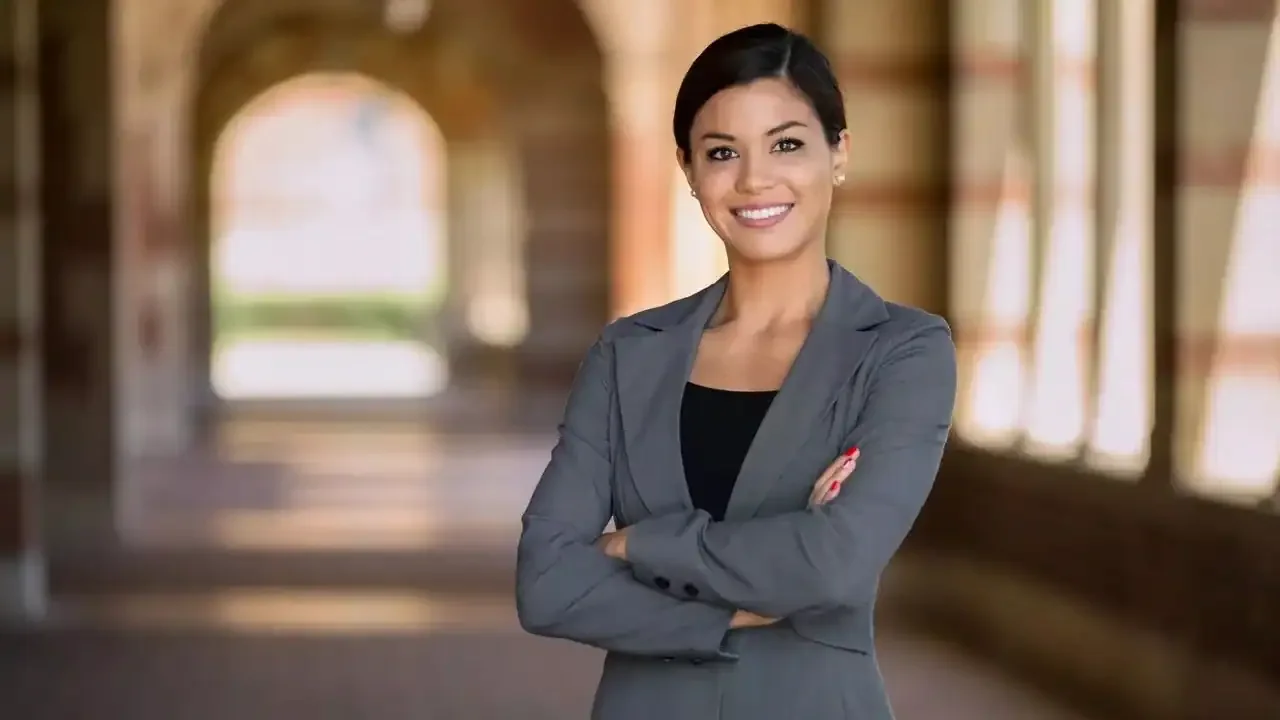 Confident woman standing with arms crossed in a blazer, smiling in a sunlit hallway.