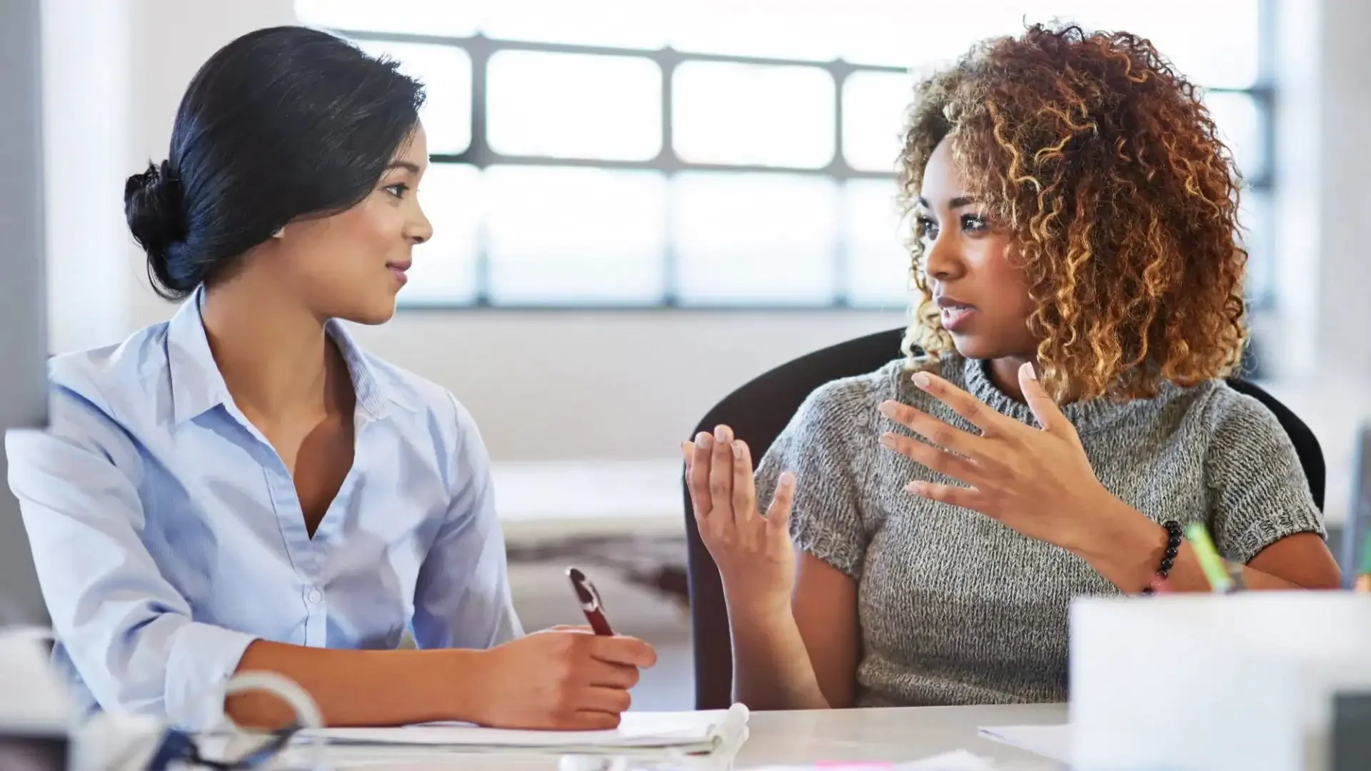 Two women having a focused conversation at work, one listening and the other speaking with hand gestures.